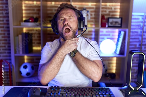 Middle age man with beard playing video games wearing headphones shouting suffocate because painful strangle. health problem. asphyxiate and suicide concept.