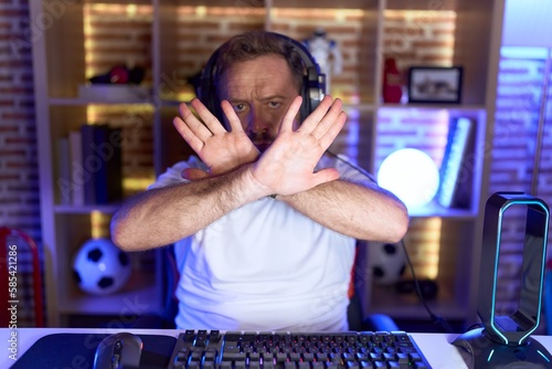 Middle age man with beard playing video games wearing headphones rejection expression crossing arms and palms doing negative sign, angry face