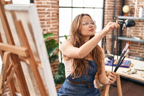 Young beautiful hispanic woman artist holding paintbrushes at art studio
