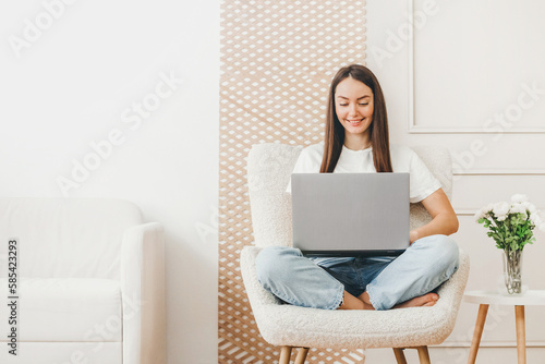 girl freelancer sits in a chair and works with a laptop at home against the background of a white wall