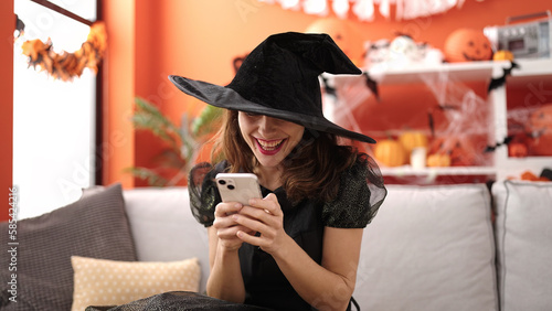 Quadro em tela Young caucasian woman wearing witch costume sitting on the sofa with smartphone