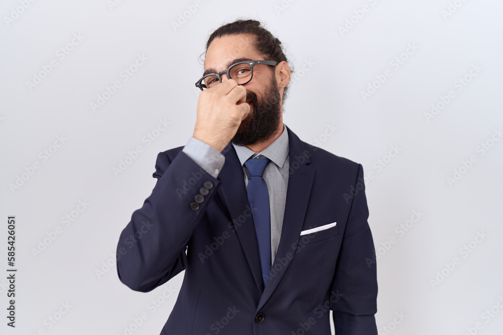 Hispanic man with beard wearing suit and tie smelling something stinky and disgusting, intolerable smell, holding breath with fingers on nose. bad smell Hispanic man with beard wearing suit and tie smelling something stinky and disgusting, intolerable smell, holding breath with fingers on nose. bad smell