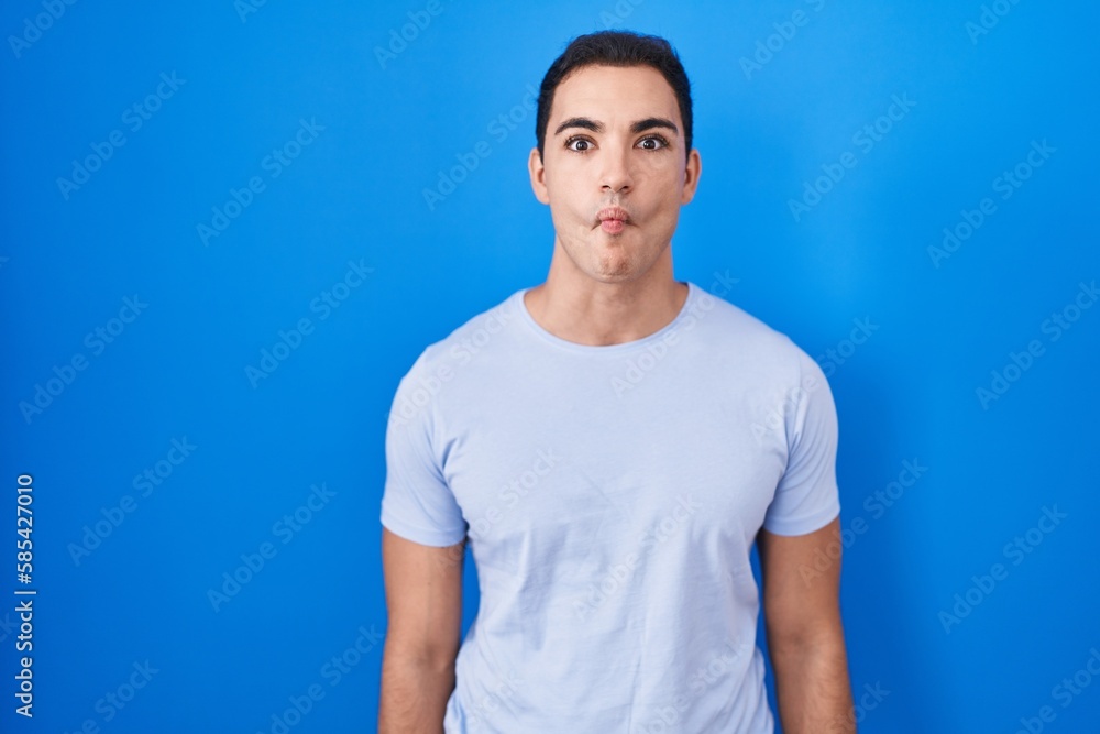 Young hispanic man standing over blue background making fish face with lips, crazy and comical gesture. funny expression. Young hispanic man standing over blue background making fish face with lips, crazy and comical gesture. funny expression.