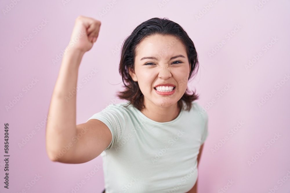 Hispanic young woman standing over pink background angry and mad raising fist frustrated and furious while shouting with anger. rage and aggressive concept. Hispanic young woman standing over pink background angry and mad raising fist frustrated and furious while shouting with anger. rage and aggressive concept.