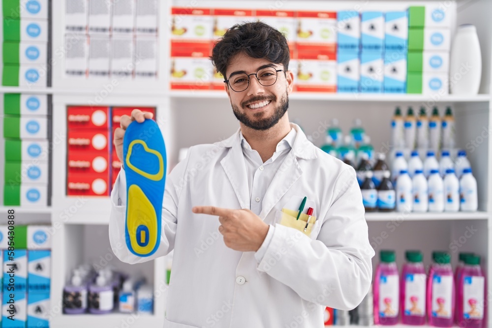 Hispanic man with beard working at pharmacy drugstore holding insole smiling happy pointing with hand and finger
