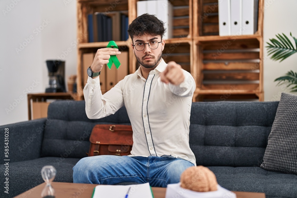 Hispanic man with beard holding support green ribbon at therapy clinic pointing with finger to the camera and to you, confident gesture looking serious Hispanic man with beard holding support green ribbon at therapy clinic pointing with finger to the camera and to you, confident gesture looking serious