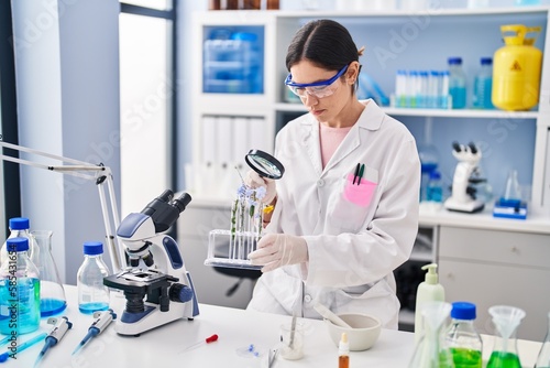 Young woman wearing scientist uniform looking flowers with magnifying glass at laboratory