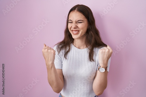 Young hispanic girl standing over pink background very happy and excited doing winner gesture with arms raised  smiling and screaming for success. celebration concept.