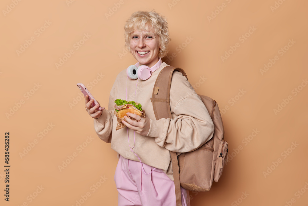 Indoor shot of cheerful blonde teenage girl uses modern cellphone for listening music and sending text messages eats burger dressed in casual clothes carries backpack isolated on brown wall. Indoor shot of cheerful blonde teenage girl uses modern cellphone for listening music and sending text messages eats burger dressed in casual clothes carries backpack isolated on brown wall.
