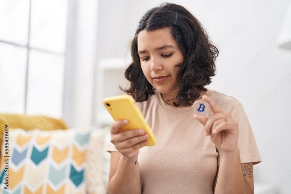 Young woman using smartphone holding bitcoin at home Young woman using smartphone holding bitcoin at home