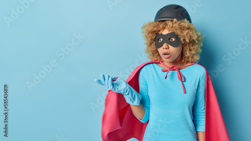 Horizontal shot of shocked confused woman with curly hair pretends having superpower dressed like hero shrugs shoulders isolated over blue background blank space for your advertising content.