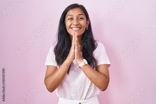 Young hispanic woman standing over pink background praying with hands together asking for forgiveness smiling confident.