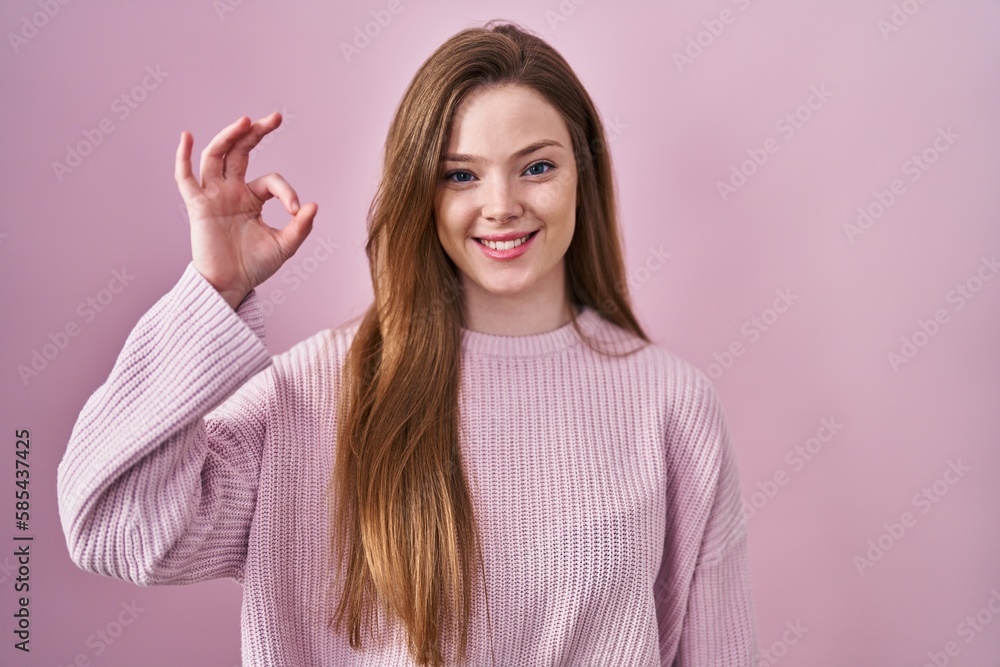 Young caucasian woman standing over pink background smiling positive doing ok sign with hand and fingers. successful expression.
