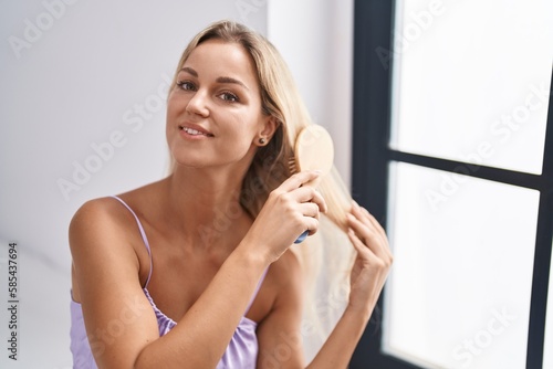 Young blonde woman combing hair sitting on bed at bedroom