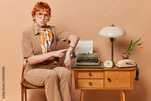 Serious strict redhead European woman indicates at watch asks not to be late dressed in elegant outfit wears big spectacles poses on chair uses retro typewriter isolated over brown background