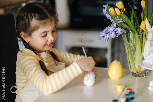 Process of little girl painting eggs on kitchen. Spring holiday. Easter decoration at home. Side view