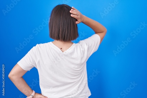 Middle age hispanic woman standing over blue background backwards thinking about doubt with hand on head