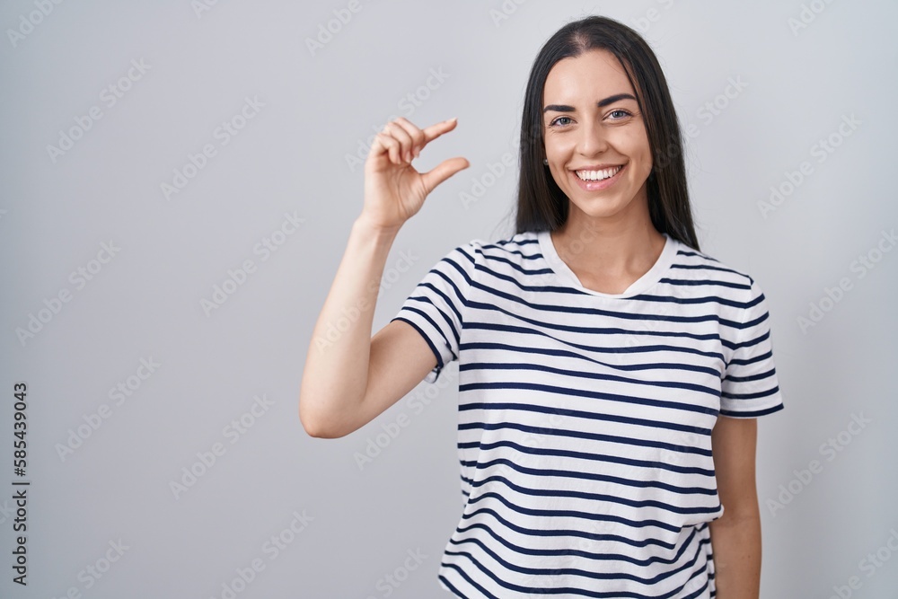 Young brunette woman wearing striped t shirt smiling and confident gesturing with hand doing small size sign with fingers looking and the camera. measure concept. Young brunette woman wearing striped t shirt smiling and confident gesturing with hand doing small size sign with fingers looking and the camera. measure concept.