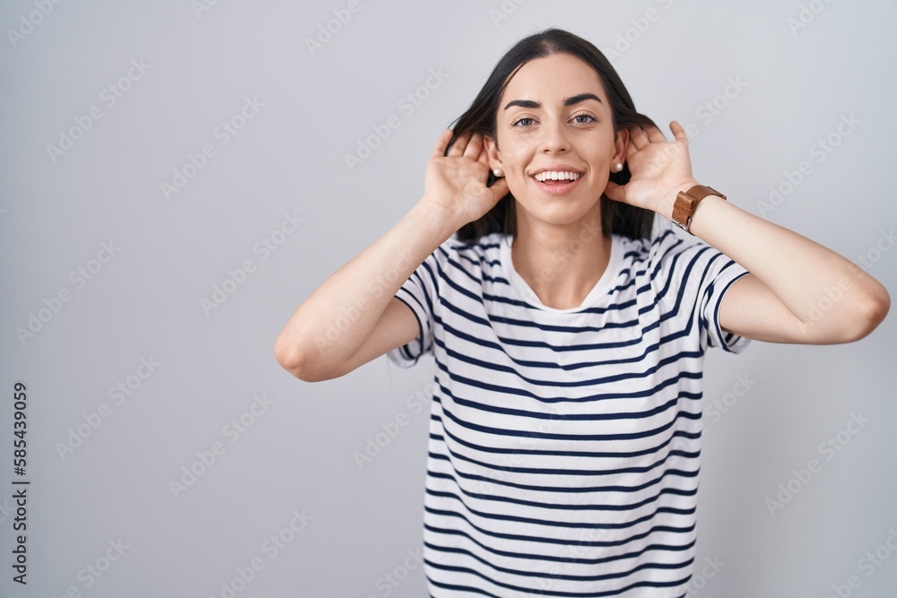 Young brunette woman wearing striped t shirt trying to hear both hands on ear gesture, curious for gossip. hearing problem, deaf Young brunette woman wearing striped t shirt trying to hear both hands on ear gesture, curious for gossip. hearing problem, deaf