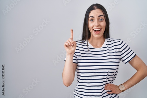 Young brunette woman wearing striped t shirt pointing finger up with successful idea. exited and happy. number one.