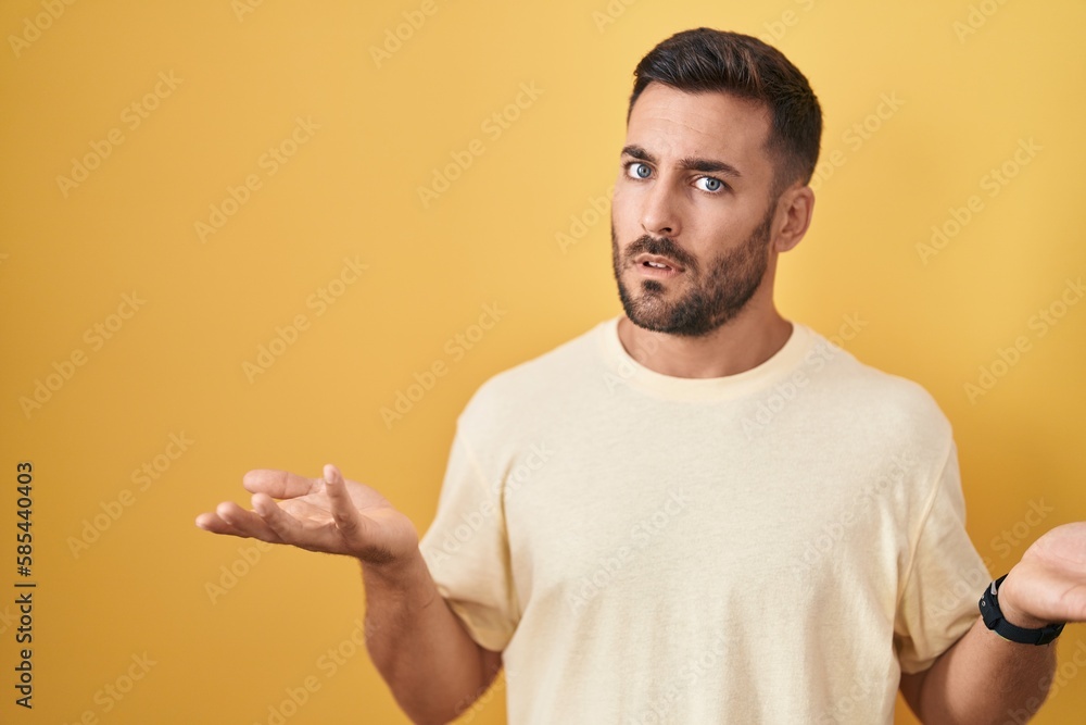 Handsome hispanic man standing over yellow background clueless and confused with open arms, no idea concept. Handsome hispanic man standing over yellow background clueless and confused with open arms, no idea concept.