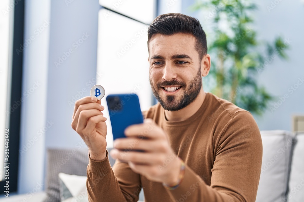 Young hispanic man using smartphone holding bitcoin at home Young hispanic man using smartphone holding bitcoin at home