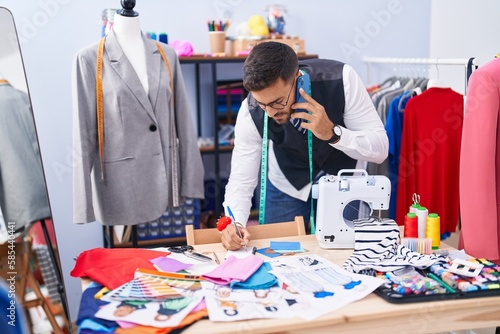 Young hispanic man tailor talking on smartphone writing on notebook at tailor shop