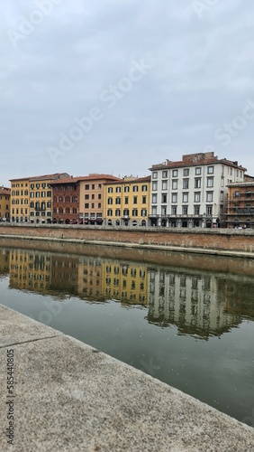 Pisa, Italy - February 25, 2023: View of the medieval town of Pisa from bridge "Ponte di Mezzo" on river Arno in winter days, with grey sky in the background. Reflection of the buildings on the water.
