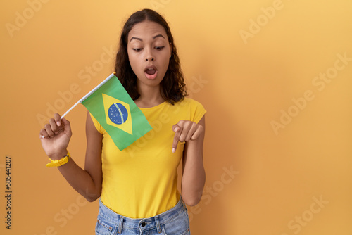 Young hispanic woman holding brazil flag pointing down with fingers showing advertisement  surprised face and open mouth