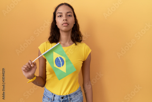 Young hispanic woman holding brazil flag relaxed with serious expression on face. simple and natural looking at the camera.