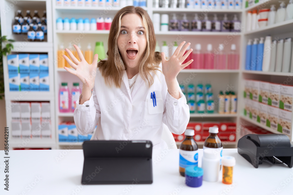 Young caucasian woman working at pharmacy drugstore using tablet celebrating victory with happy smile and winner expression with raised hands