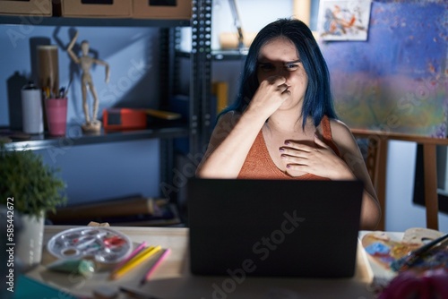 Young modern girl with blue hair sitting at art studio with laptop at night smelling something stinky and disgusting  intolerable smell  holding breath with fingers on nose. bad smell