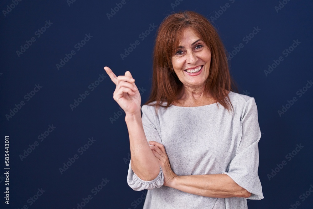 Middle age woman standing over blue background with a big smile on face, pointing with hand and finger to the side looking at the camera. Middle age woman standing over blue background with a big smile on face, pointing with hand and finger to the side looking at the camera.