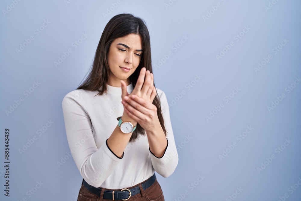 Young brunette woman standing over blue background suffering pain on hands and fingers, arthritis inflammation Young brunette woman standing over blue background suffering pain on hands and fingers, arthritis inflammation