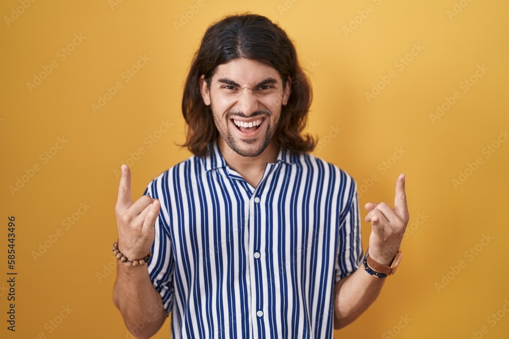 Hispanic man with long hair standing over yellow background shouting with crazy expression doing rock symbol with hands up. music star. heavy concept. Hispanic man with long hair standing over yellow background shouting with crazy expression doing rock symbol with hands up. music star. heavy concept.