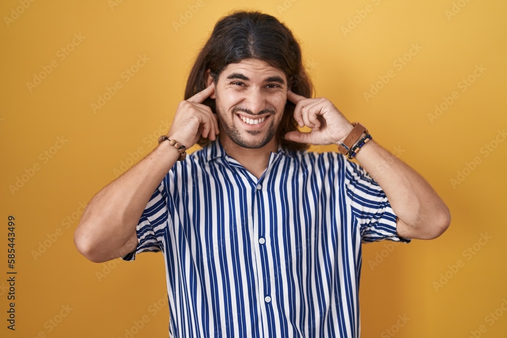Hispanic man with long hair standing over yellow background covering ears with fingers with annoyed expression for the noise of loud music. deaf concept. Hispanic man with long hair standing over yellow background covering ears with fingers with annoyed expression for the noise of loud music. deaf concept.