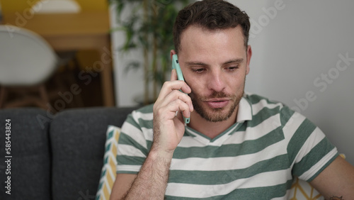Young caucasian man speaking on the phone sitting on the sofa at home