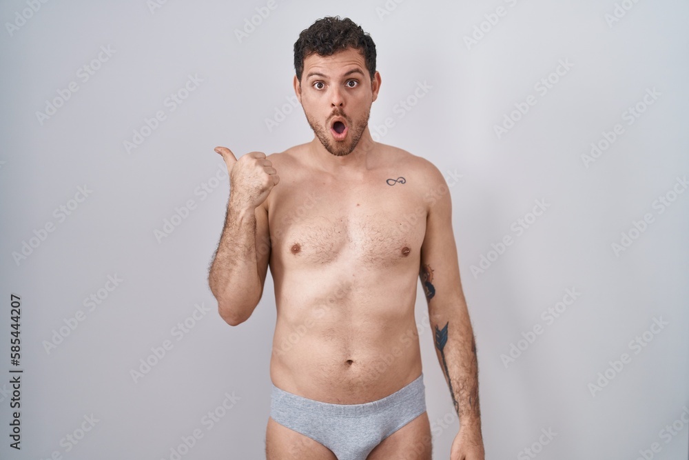Young hispanic man standing shirtless wearing underware surprised pointing with hand finger to the side, open mouth amazed expression.