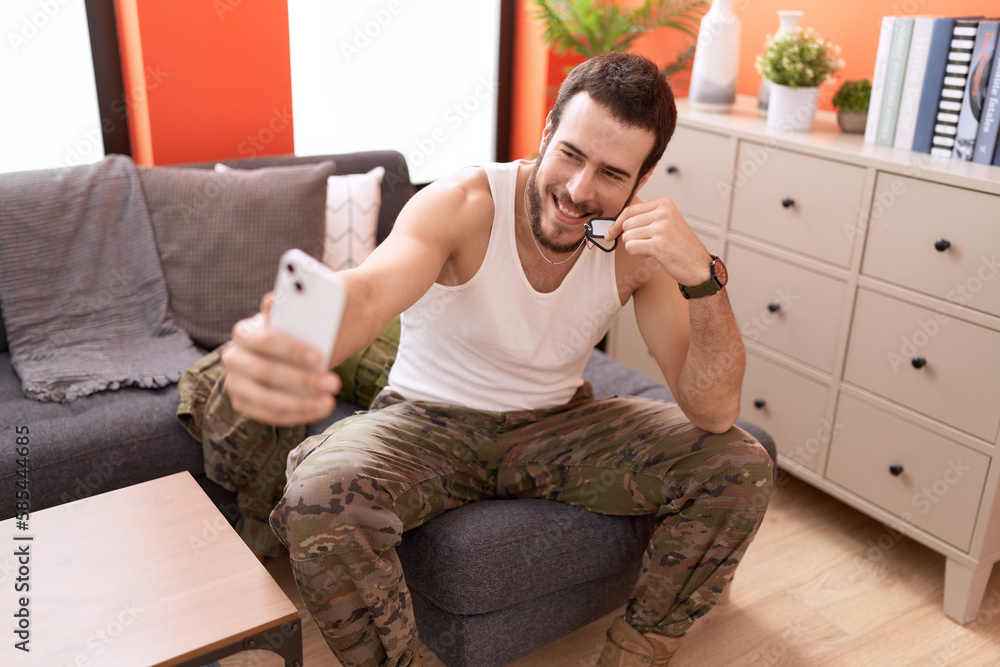 Fototapeta premium Young hispanic man wearing soldier uniform make selfie by smartphone at home