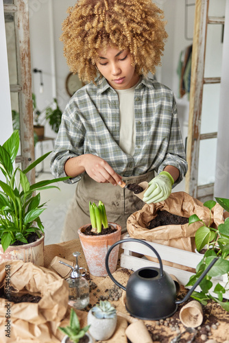 Spring hobby concept. Curly haired female botanist holds small pot involved in gardening wears checkered shirt apron and protective glove takes care of houseplant stands near table with soil
