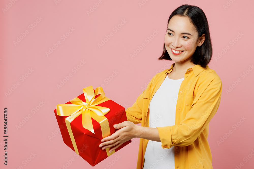 Young fun woman of Asian ethnicity wear yellow shirt white t-shirt hold in hand give present box with gift ribbon bow isolated on plain pastel light pink background studio portrait. Lifestyle concept. Young fun woman of Asian ethnicity wear yellow shirt white t-shirt hold in hand give present box with gift ribbon bow isolated on plain pastel light pink background studio portrait. Lifestyle concept.