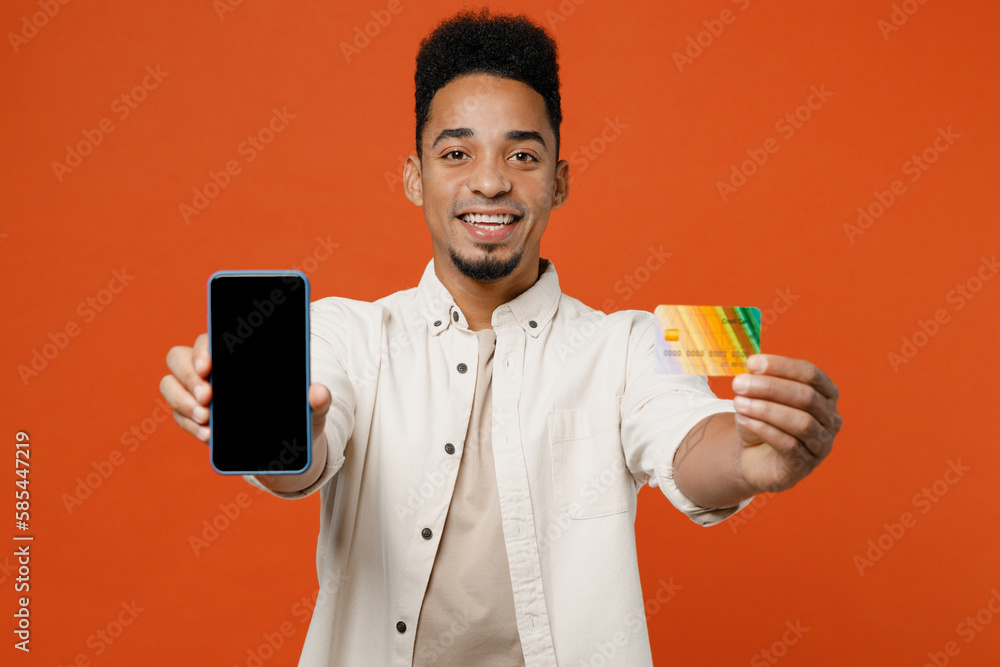 Young man of African American ethnicity wear light casual clothes using blank screen mobile cell phone hold credit bank card shopping online order delivery book tour isolated on orange red background. Young man of African American ethnicity wear light casual clothes using blank screen mobile cell phone hold credit bank card shopping online order delivery book tour isolated on orange red background.