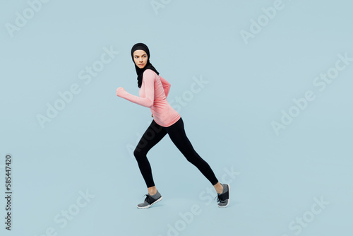 Full body young asian fitness trainer sporty woman in pink abaya hijab spend time in gym stand at start going to run look aside isolated on plain blue background studio Workout sport fit abs concept