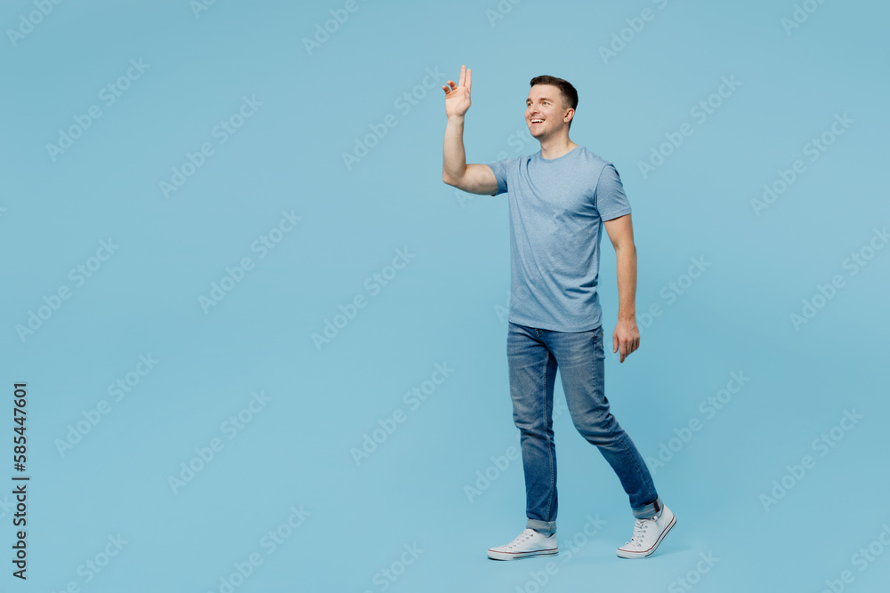 Full body side view young smiling happy man wears casual t-shirt walking strolling going waving hand greet meet isolated on plain pastel light blue cyan background studio portrait. Lifestyle concept. Full body side view young smiling happy man wears casual t-shirt walking strolling going waving hand greet meet isolated on plain pastel light blue cyan background studio portrait. Lifestyle concept.