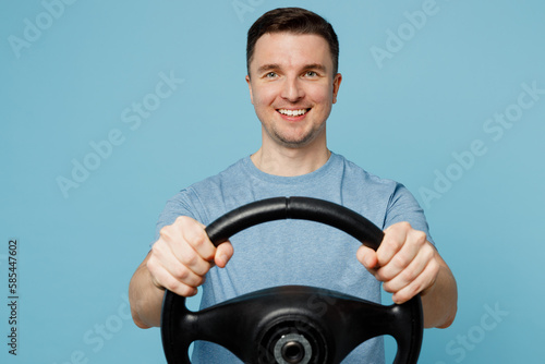 Young satisfied cheerful smiling fun happy man wear casual t-shirt hold steering wheel driving car look camera isolated on plain pastel light blue cyan background studio portrait. Lifestyle concept.