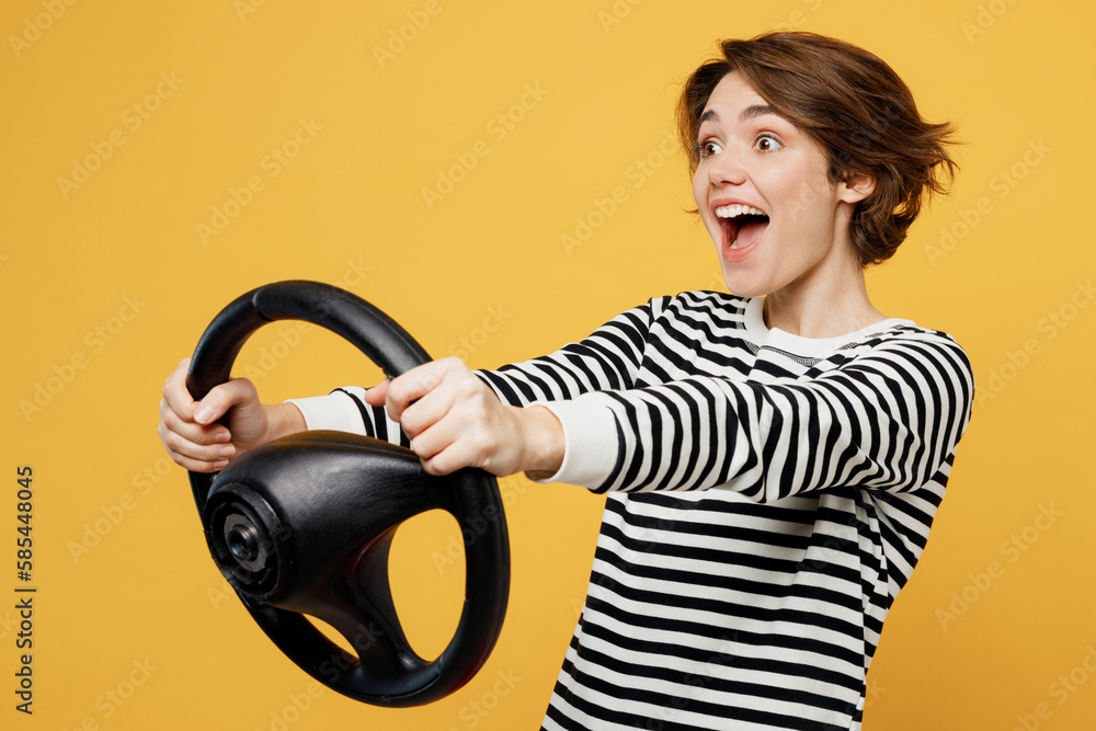 Young surprised happy fun cool woman wear casual striped black and white shirt hold steering wheel driving car look camera isolated on plain yellow color background studio portrait. Lifestyle concept.