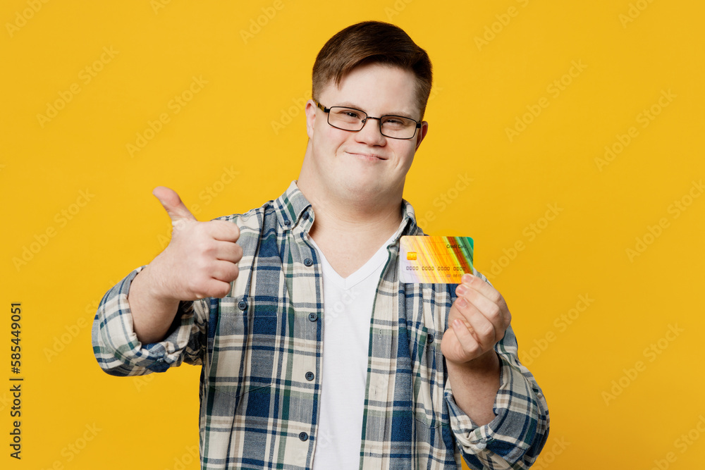 Young smiling man with down syndrome wear glasses casual clothes hold credit bank card show thumb up like gesture isolated on pastel plain yellow color background. Genetic disease world day concept. Young smiling man with down syndrome wear glasses casual clothes hold credit bank card show thumb up like gesture isolated on pastel plain yellow color background. Genetic disease world day concept.