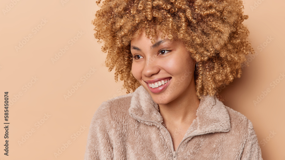 Headshot of pleased radiant woman with curly hair smiles toothily has dreamy satisfied expression dressed in warm fur jacket isolated over brown background copy space for your promotional content Headshot of pleased radiant woman with curly hair smiles toothily has dreamy satisfied expression dressed in warm fur jacket isolated over brown background copy space for your promotional content