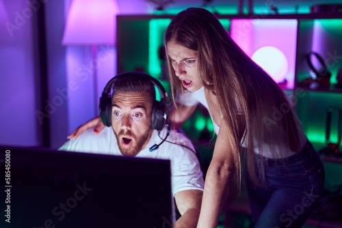 Young couple playing video games in shock face, looking skeptical and sarcastic, surprised with open mouth