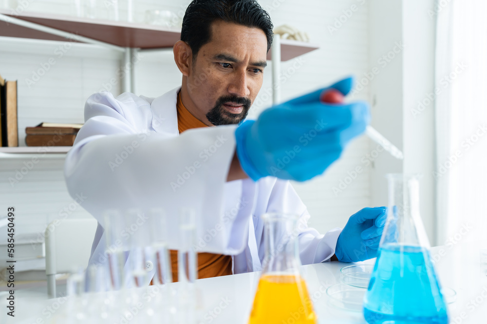 A male teacher in white lab coat with blue rubber gloves with many laboratory tools on shelves and table. Using pipette at right hand drop chemical fluids in blue chemical flask on table carefully. A male teacher in white lab coat with blue rubber gloves with many laboratory tools on shelves and table. Using pipette at right hand drop chemical fluids in blue chemical flask on table carefully.